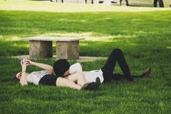 Man And Woman Laying On Green Grass Near Concrete Bench